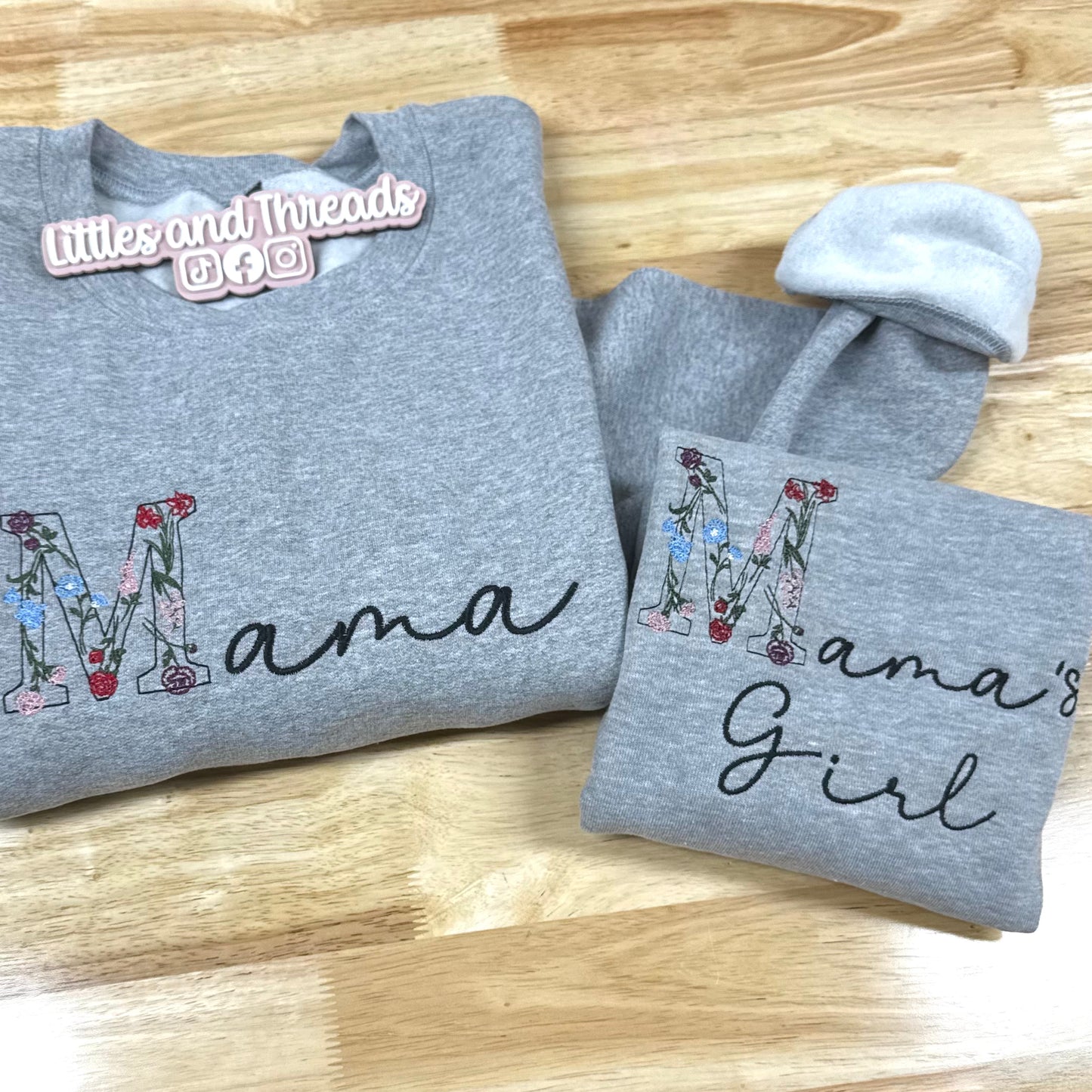 Gray crewneck with 'Mama' and 'Mama's Girl' text on a wooden surface, featuring the brand 'littles and threads'.