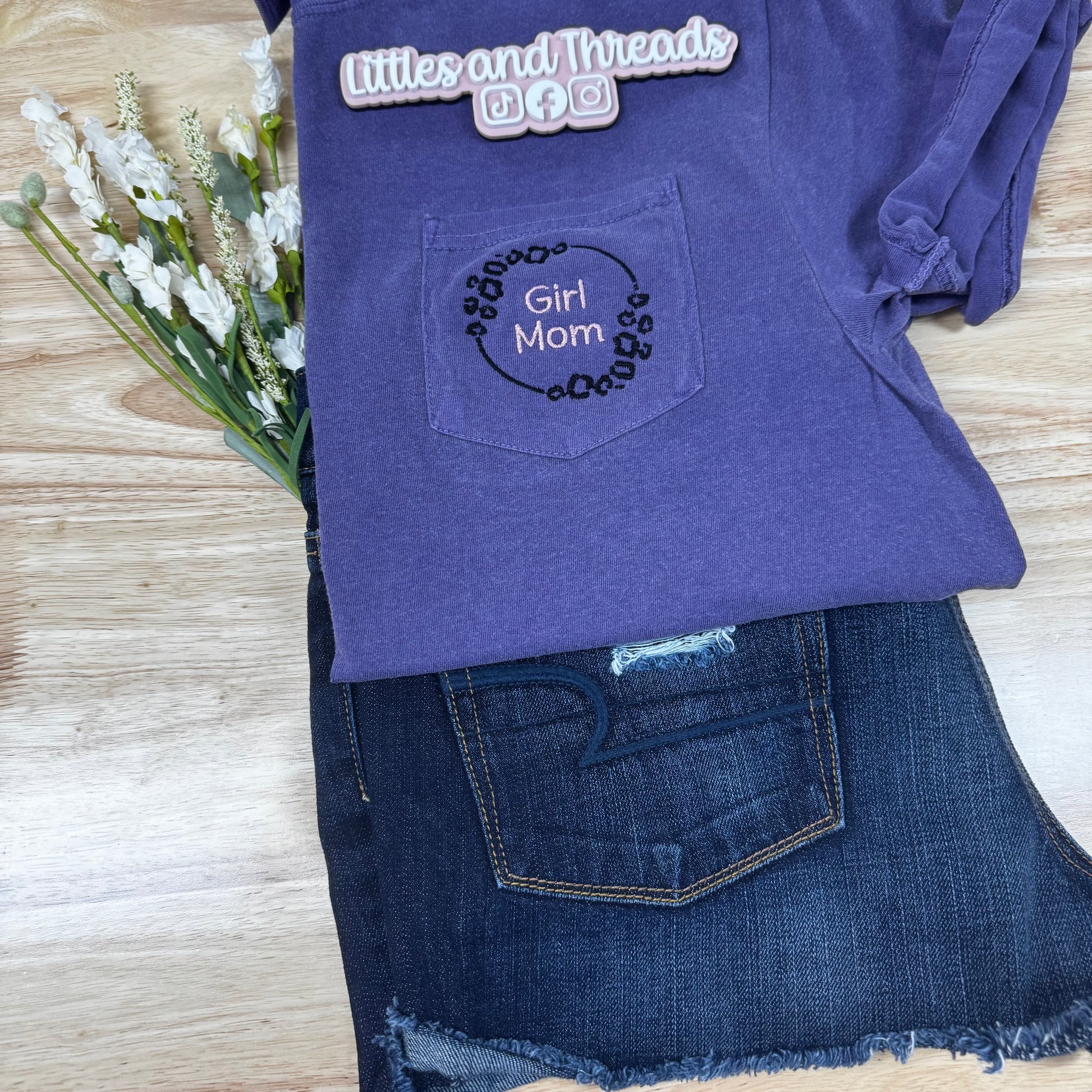 Purple shirt with 'Girl Mom' embroidery on a wooden surface with flowers.