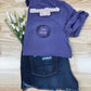 Purple shirt with 'Girl Mom' design on a wooden surface with denim shorts and flowers.