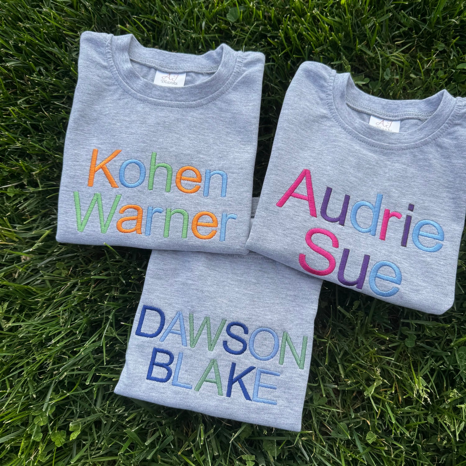 Three folded gray shirts with embroidered names on a grass background
