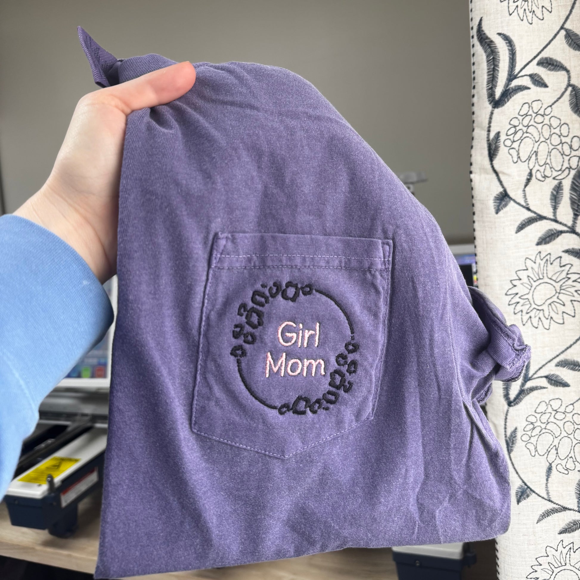 Purple shirt with 'Girl Mom' embroidery held by a person in an office setting.