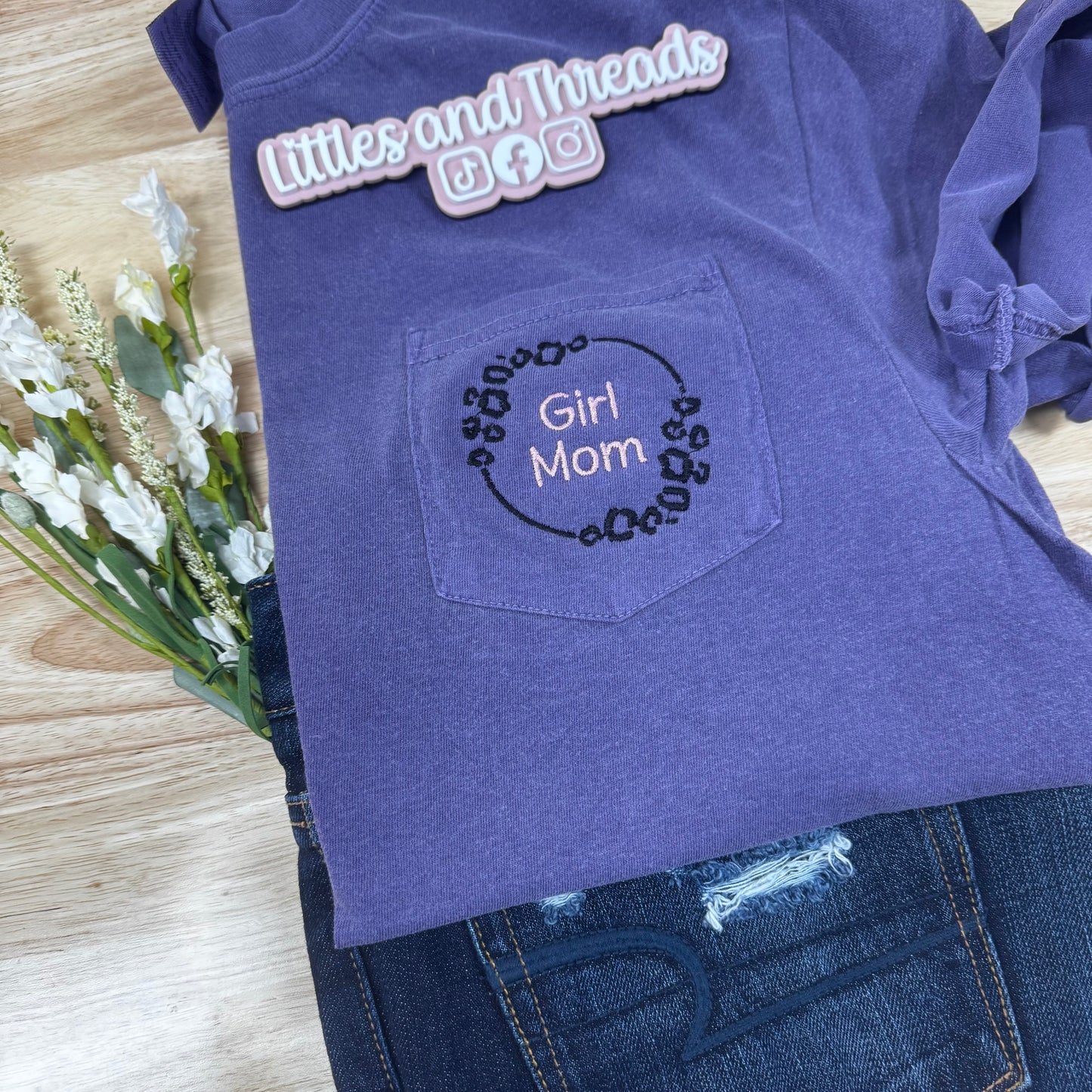 Purple t-shirt with 'Girl Mom' design on a wooden surface with flowers.