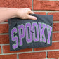 Hand holding a black t-shirt with 'SPooky' in purple letters against a brick wall.