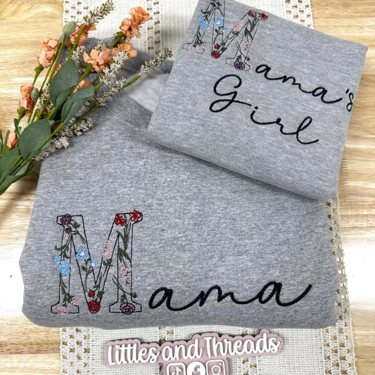 Gray sweatshirt with 'Mama's Girl' and 'Mama' text on a wooden surface with flowers.